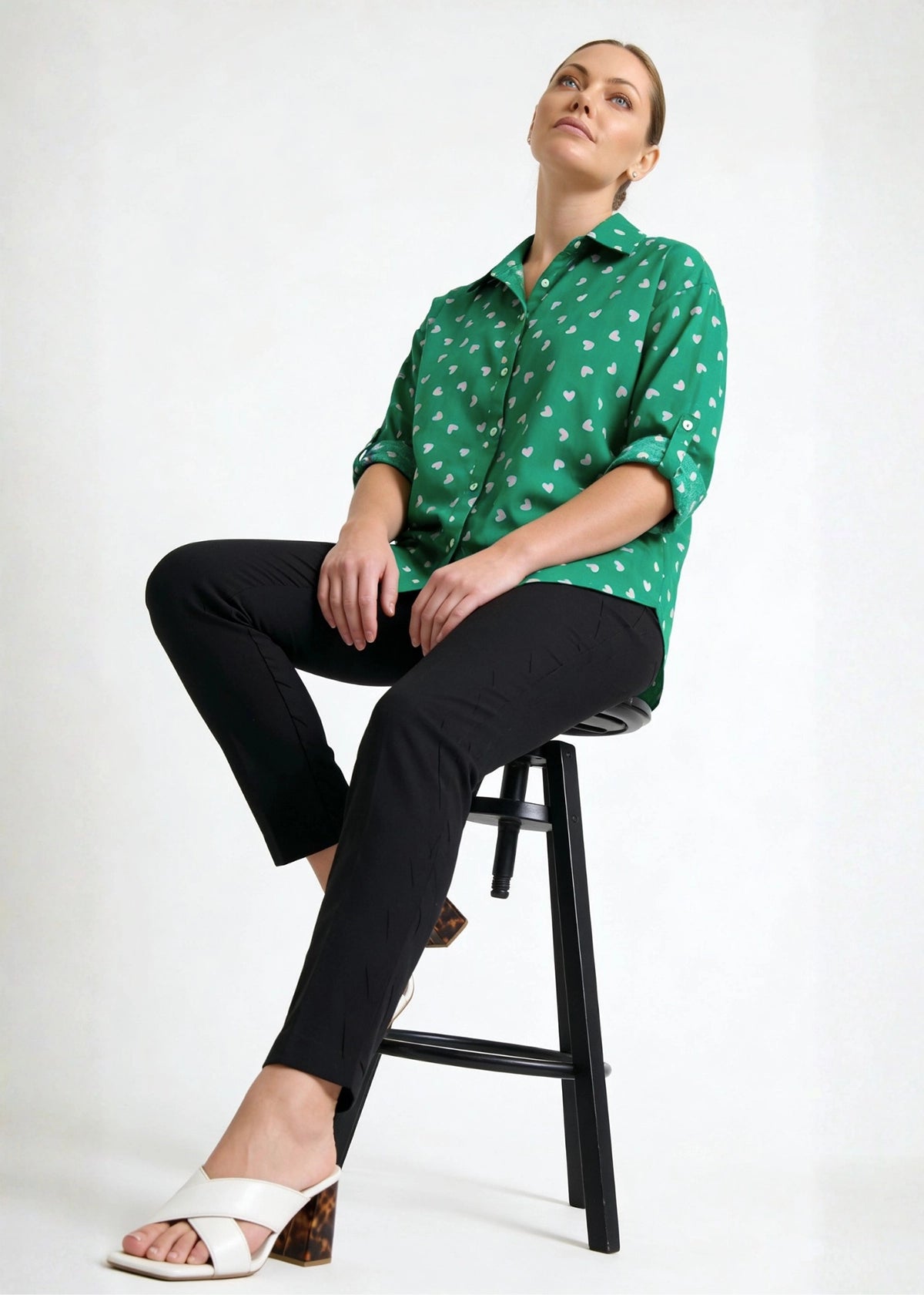 Person wearing a green patterned shirt and black pants sitting on a stool against a white background